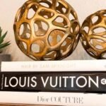 Two gold sculptures sit atop a stack of books, accompanied by a small green plant, creating an elegant display.