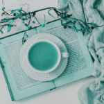 Open book with cup of tea stacked on top of it surrounded by soft knit fabric and floral stems