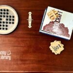 On a wooden table, a stack of books, a stack of coasters, and two wooden games are artfully arranged.
