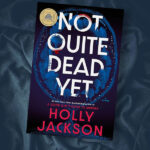 Flatlay of the book Not Quite Dead Yet by Holly Jackson on a dark rumpled fabric covered background