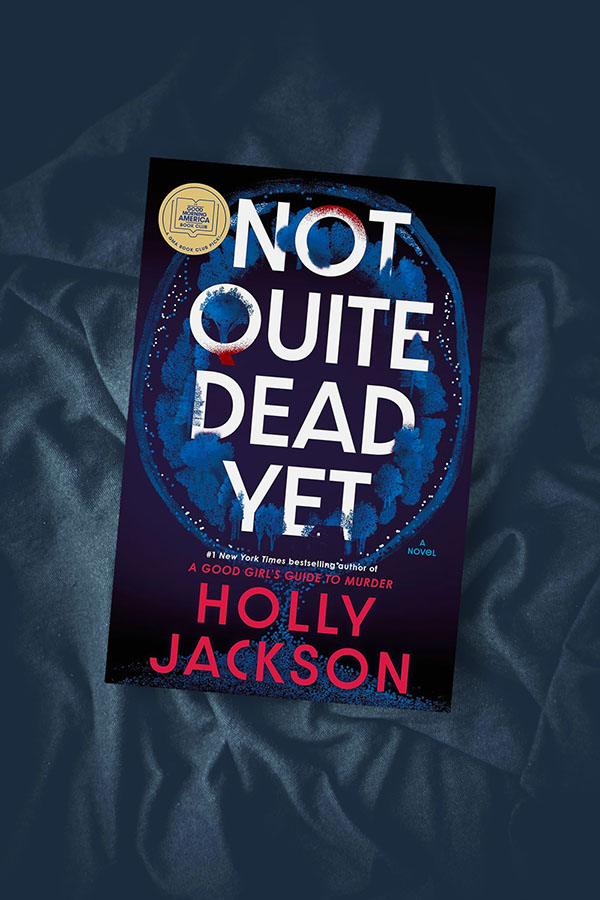 Flatlay of the book Not Quite Dead Yet by Holly Jackson on a dark rumpled fabric covered background