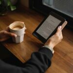 Hangs holding a coffee mug and an ereader at a table near a window