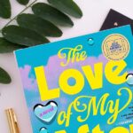 Flatlay of The Love of My Afterlife by Kirsty Greenwood with a plant, a notebook, and a pen